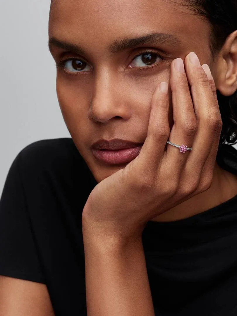 Nahaufnahme des rosa Blumenrings am Ringfinger, Hand am Gesicht abgestützt, neutraler Hintergrund.