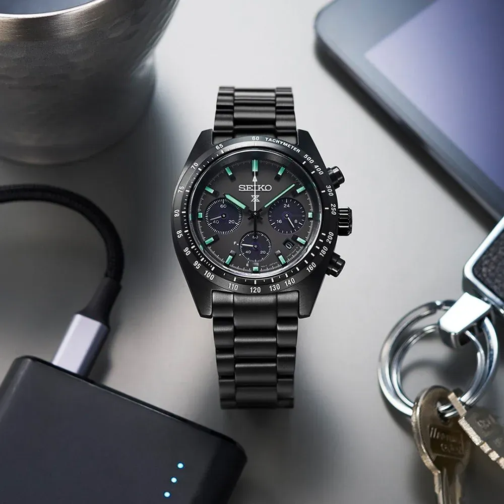 Vue de dessus de la montre entourée d'accessoires comme des clés et des appareils électroniques sur fond gris clair.