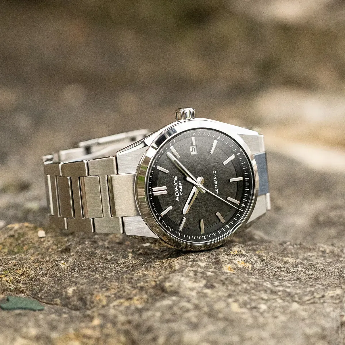Reloj apoyado sobre una superficie de roca rugosa, toma en ángulo diagonal con fondo natural desenfocado.