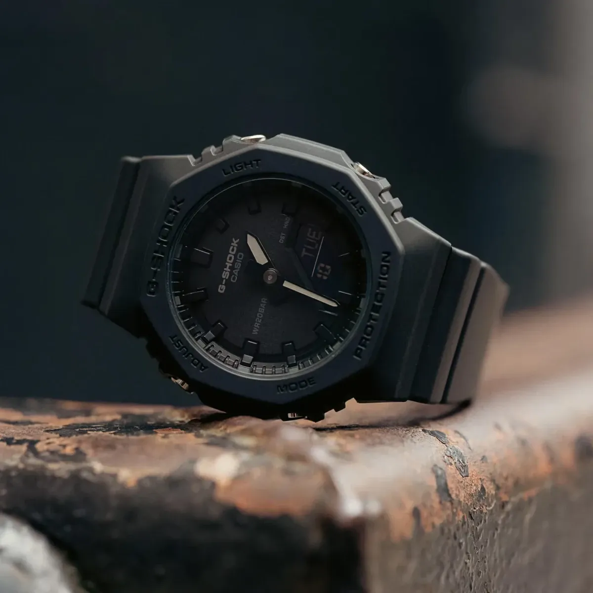 Reloj negro apoyado sobre una superficie de metal oxidado, toma en ángulo con fondo oscuro desenfocado.