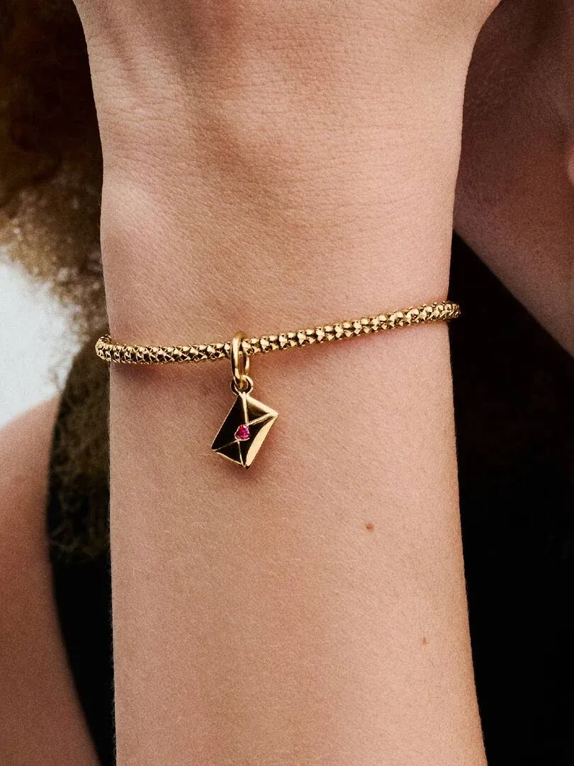 Gros plan d'un charm enveloppe doré avec zirconie rouge sur un bracelet, porté au poignet.