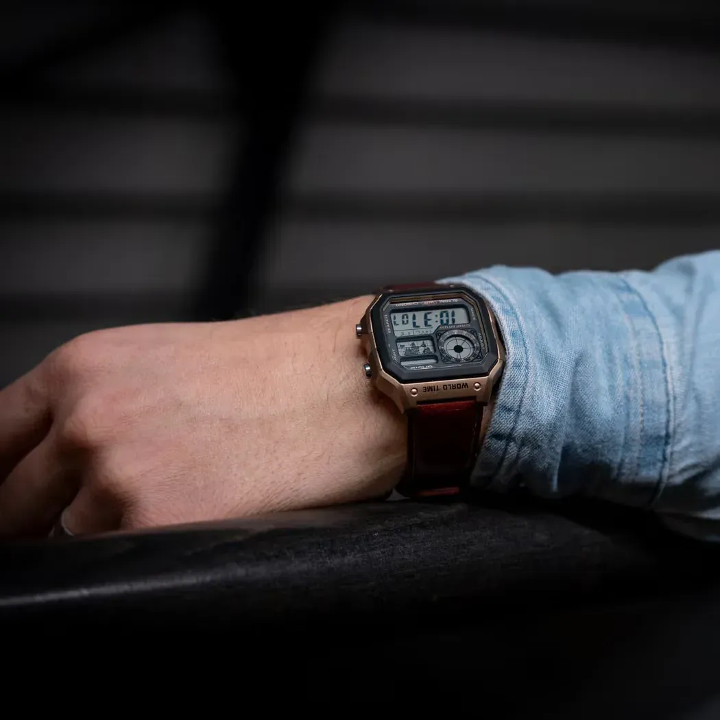 Primer plano del reloj puesto en la muñeca, brazo apoyado sobre superficie oscura, fondo urbano desenfocado.