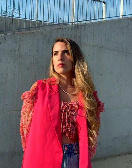 Model met parelsnoer, roze blazer en bloemprint top, poserend voor een betonnen muur buiten.