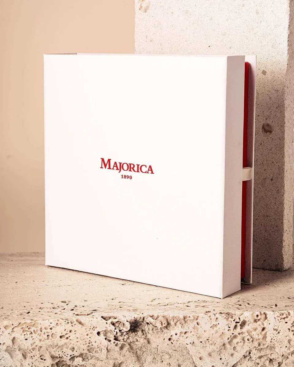 Boîte blanche de la marque Majorica avec logo rouge, vue de face en angle sur une surface en pierre claire.