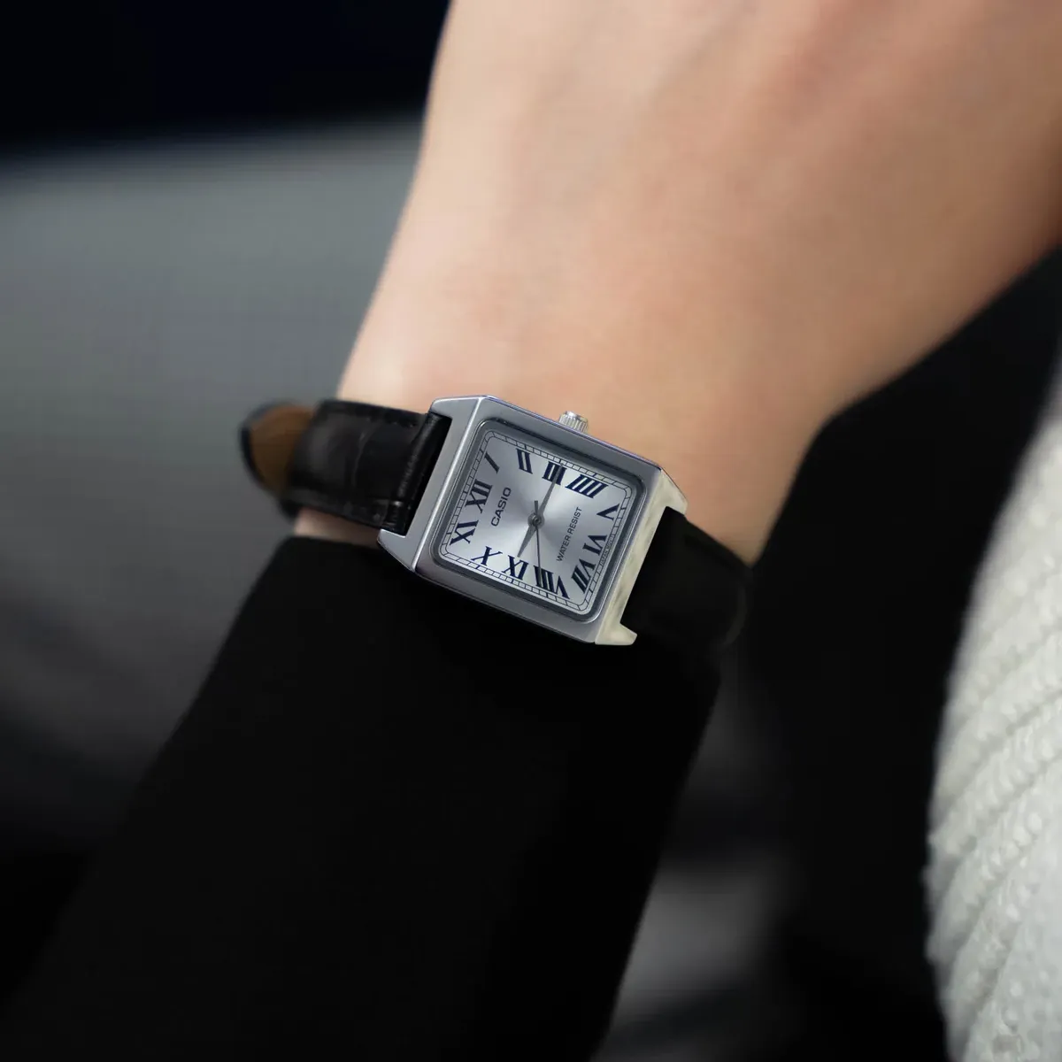 Reloj cuadrado puesto en la muñeca, toma en ángulo, fondo desenfocado con ropa en negro y blanco.
