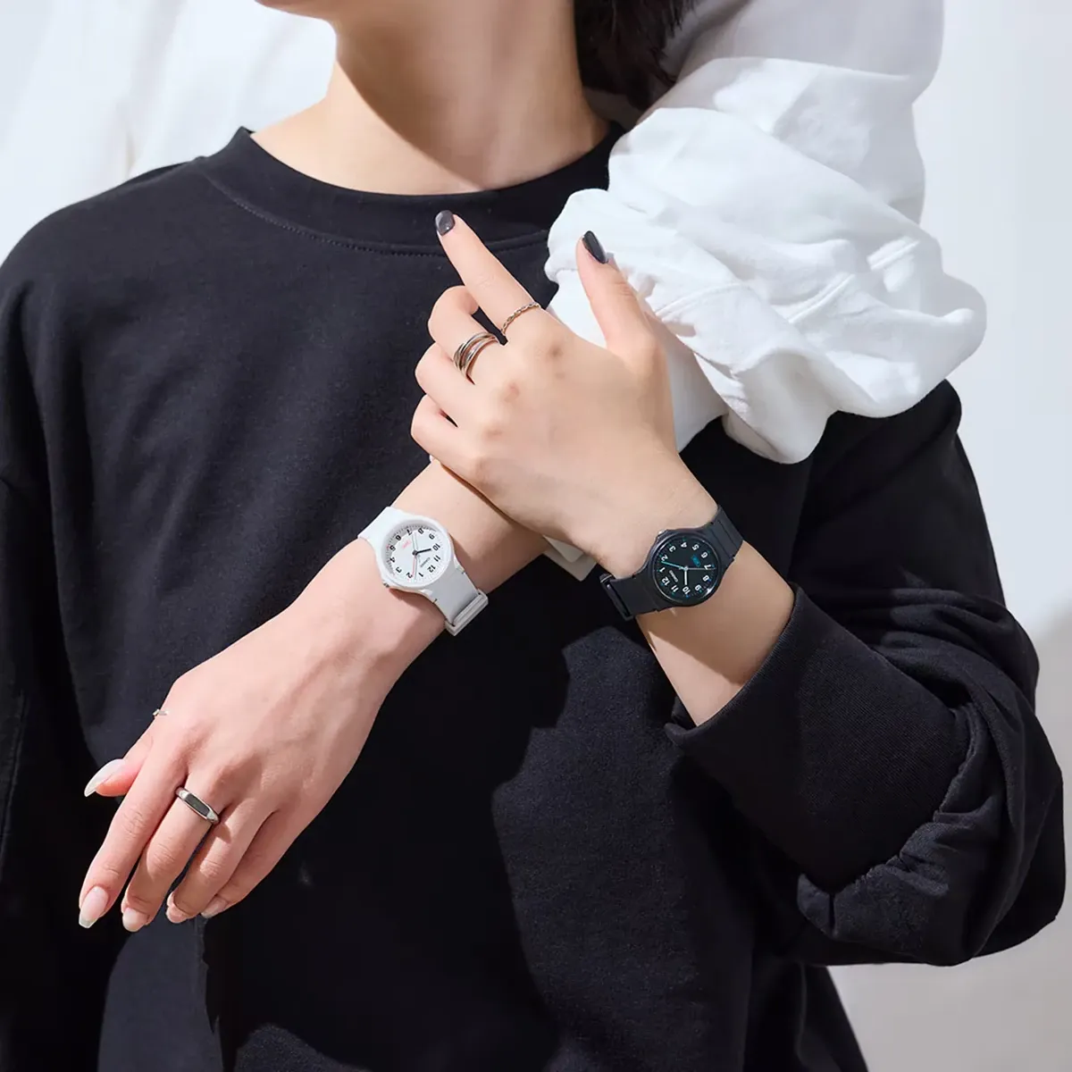 Plano medio de muñecas con relojes blanco y negro puestos, modelo con ropa negra sobre fondo neutro.