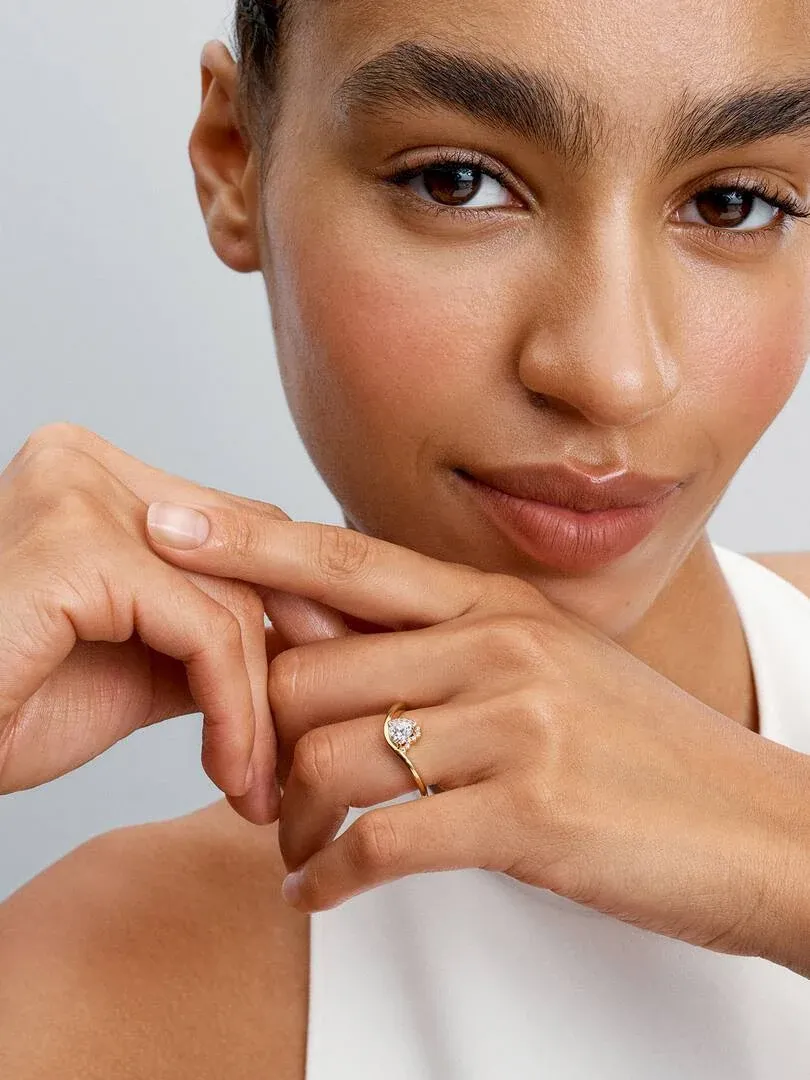 Gros plan de la bague cœur dorée portée au doigt, avec le visage d'un modèle flou en arrière-plan.