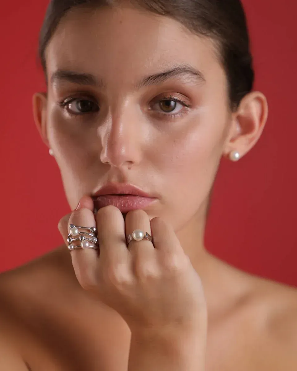 Retrato de modelo luciendo anillos de perlas y pendientes sobre fondo rojo sólido.