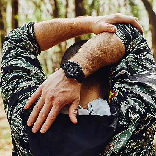 Reloj puesto en la muñeca de un modelo con chaqueta de camuflaje, fondo de bosque desenfocado.