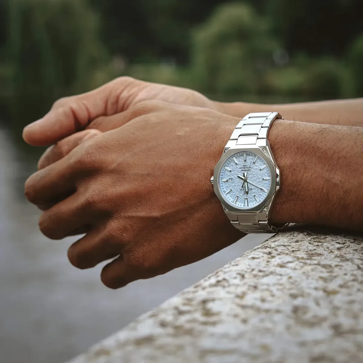 Reloj puesto en la muñeca, brazos apoyados sobre un murete de piedra con fondo natural desenfocado.