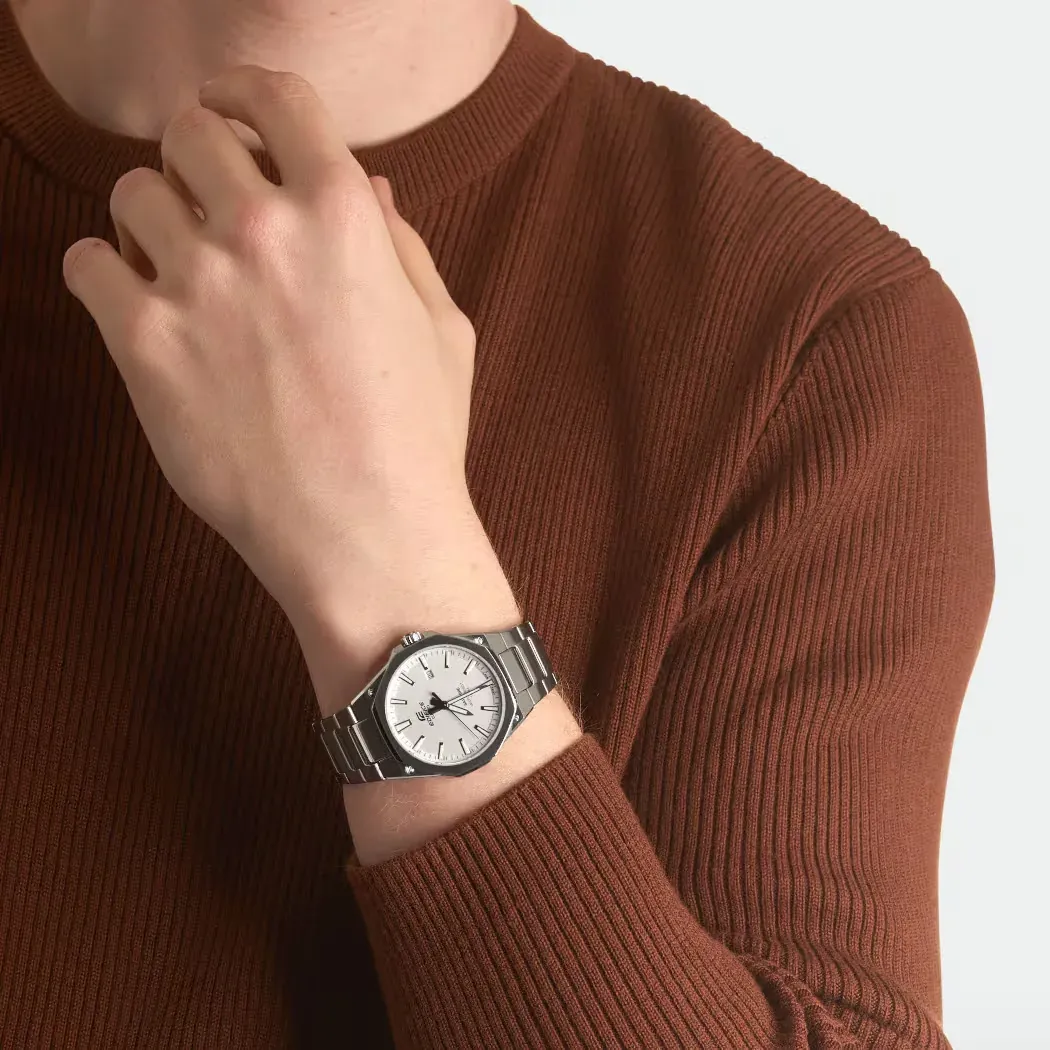 Reloj puesto en la muñeca de un modelo con jersey marrón acanalado, fondo neutro, toma de plano medio.