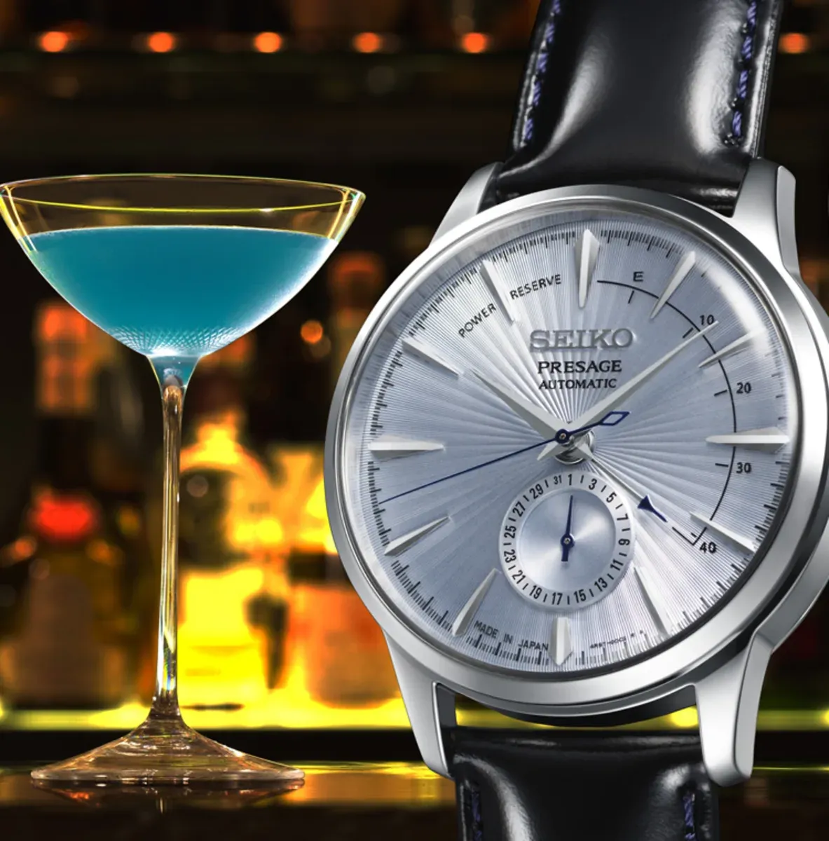 Watch next to a blue cocktail glass, blurred bar background with warm lighting, still life shot.