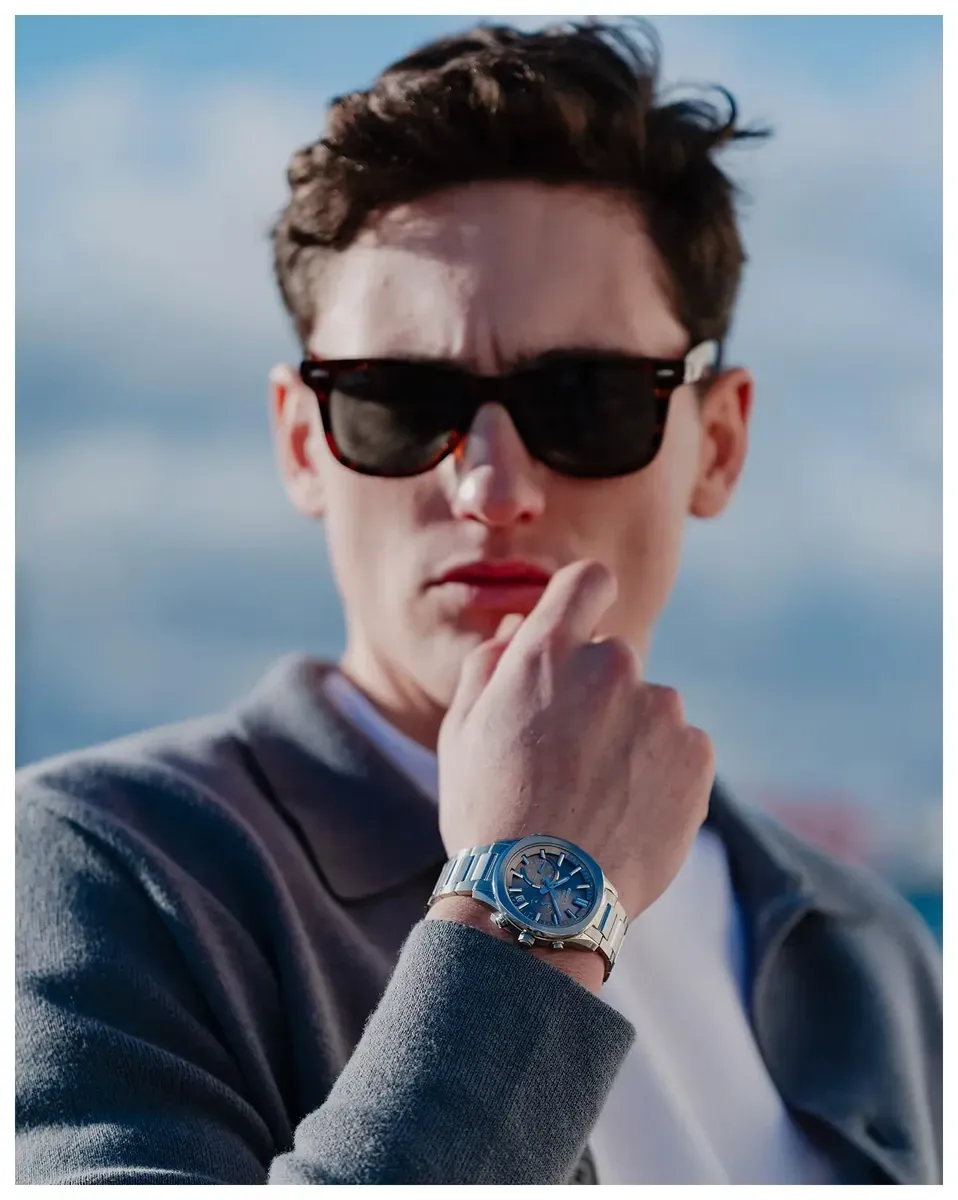 Modelo masculino con gafas de sol y chaqueta gris, reloj puesto en la muñeca, fondo exterior luminoso desenfocado.