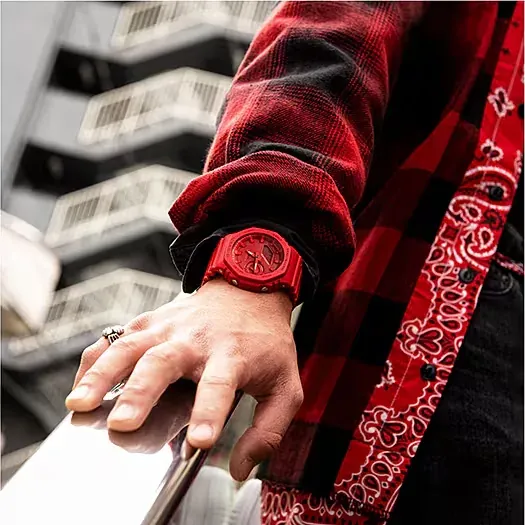 Primer plano del reloj rojo puesto en la muñeca, mano apoyada sobre superficie reflectante, fondo urbano desenfocado.