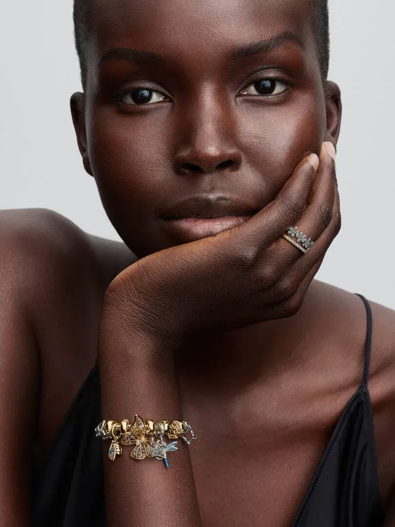 Nahaufnahme von Hand und Handgelenk mit einem Charm-Armband und Ring vor hellem, neutralem Hintergrund.