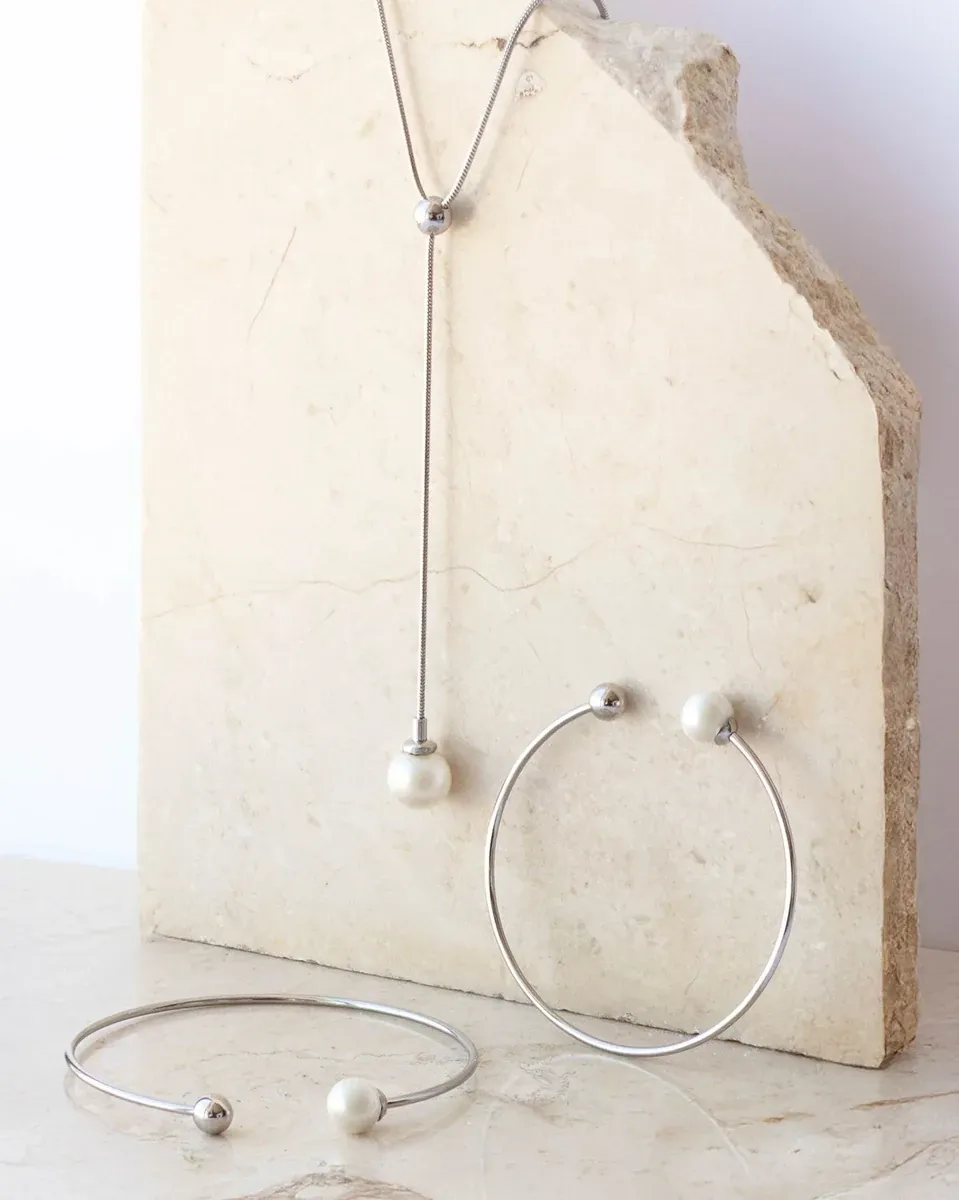 Collier et bracelet en acier avec perles blanches sur un socle en pierre naturelle claire, composition artistique.