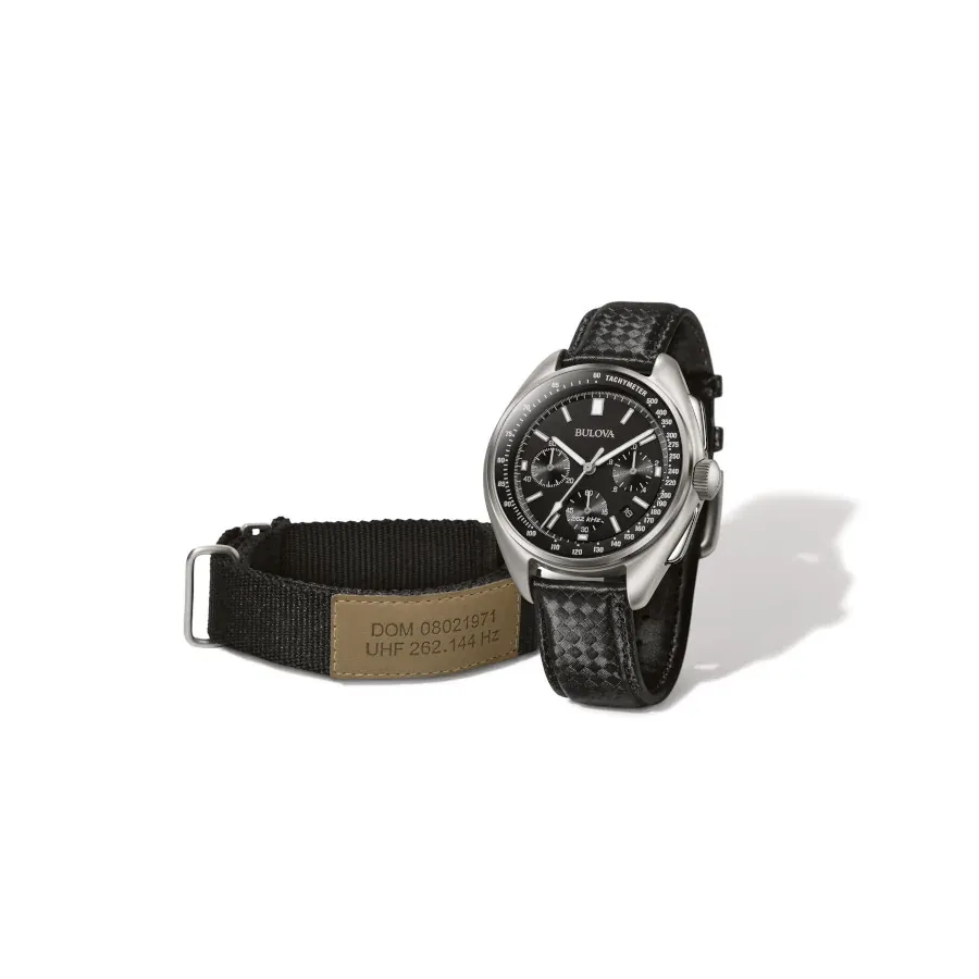 Reloj en posición vertical angulada junto a una correa textil negra con etiqueta técnica, fondo blanco.