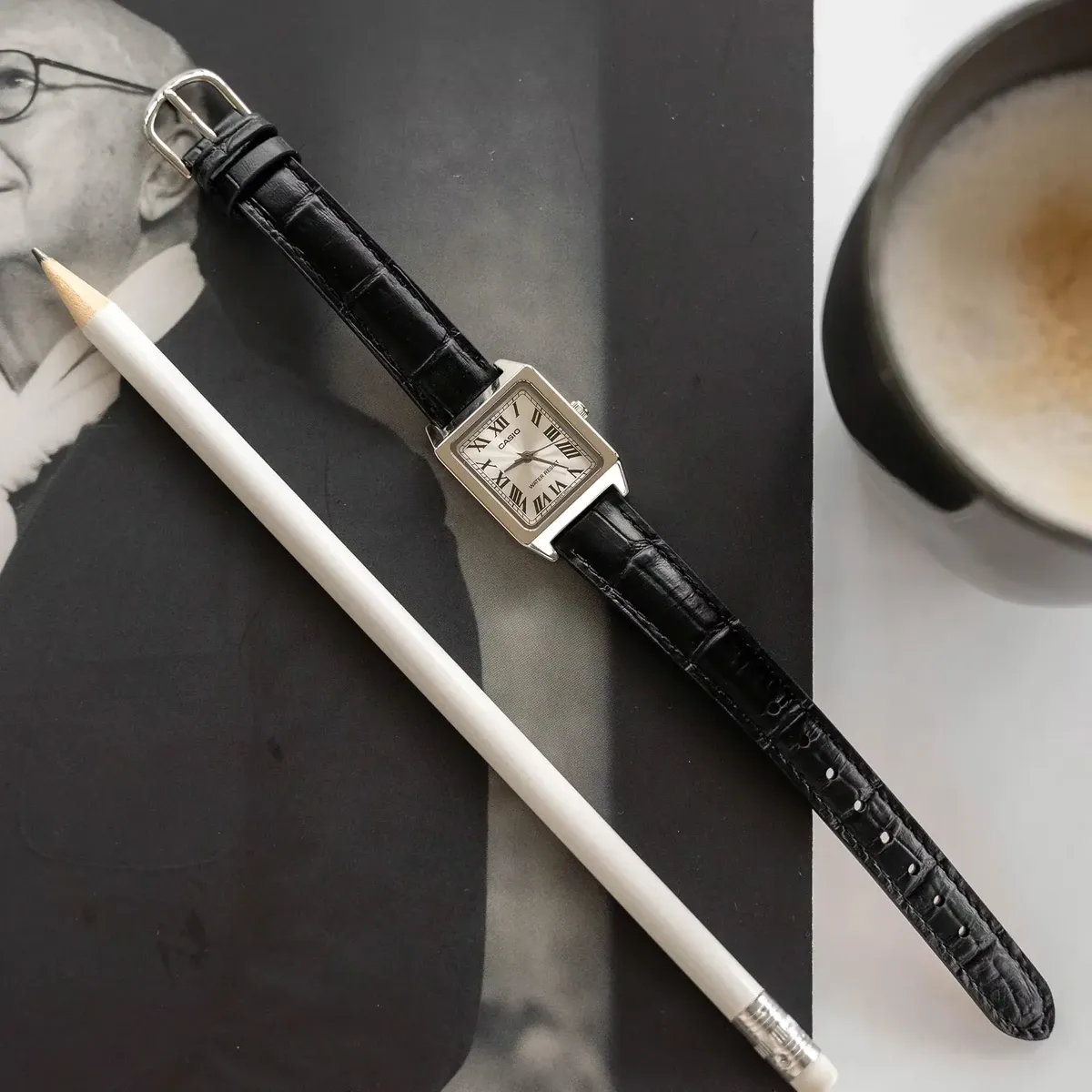 Reloj cuadrado en plano cenital sobre fotografía en blanco y negro, acompañado de un lápiz blanco y una taza de café.
