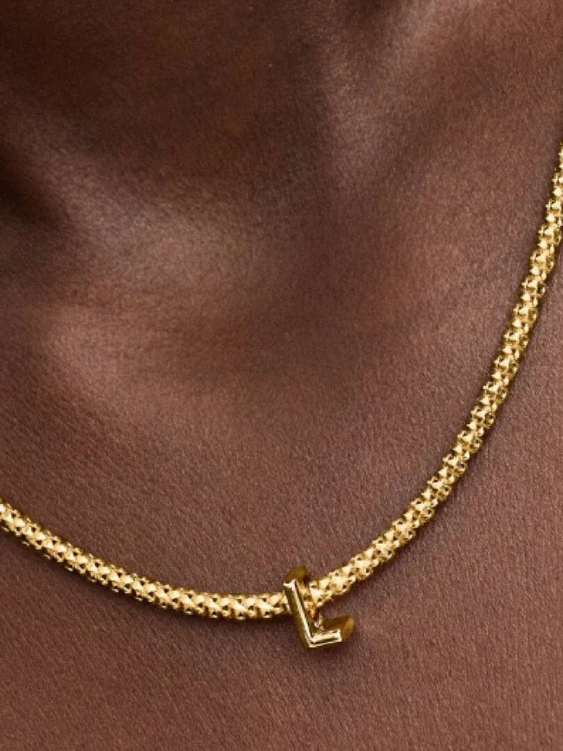 Nahaufnahme des Buchstaben L Charms an einer goldenen Halskette am Hals eines Models, Seitenansicht.