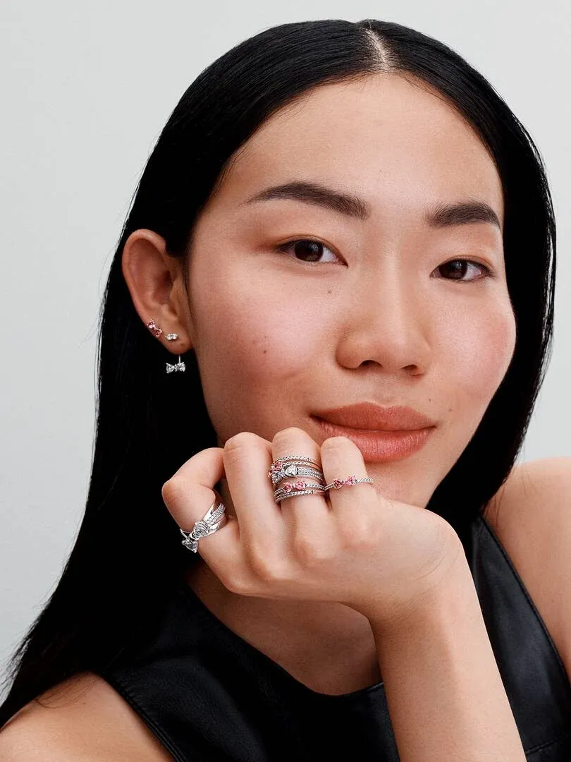 Close-up of a model wearing pink bow stud earrings and multiple silver rings, light neutral background.