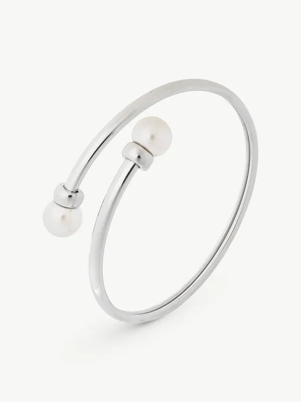 Brazalete rígido de acero con dos perlas blancas en los extremos, vista en ángulo sobre fondo blanco.