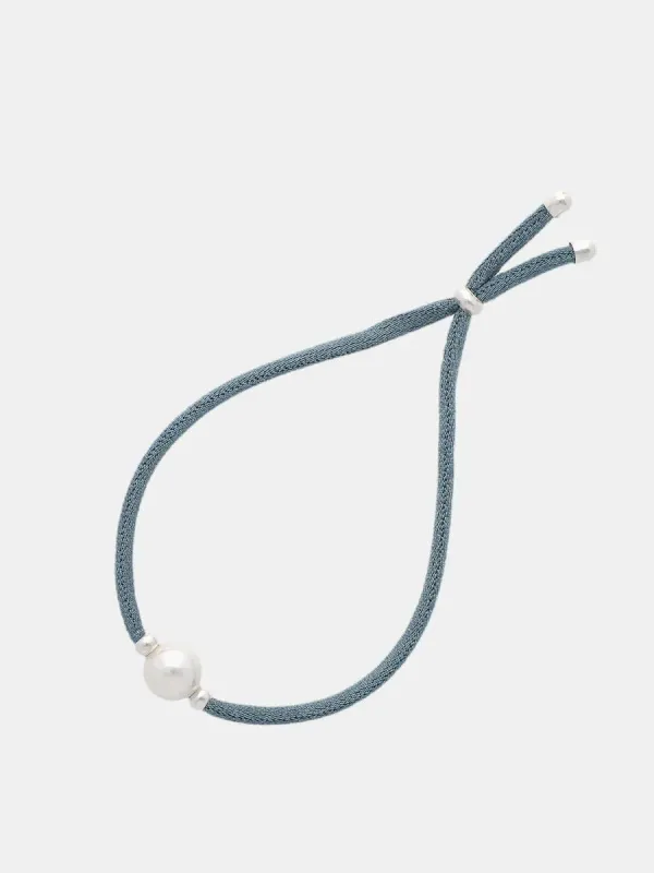 Pulsera de cordón azul con una perla blanca central, vista cenital sobre fondo gris claro.