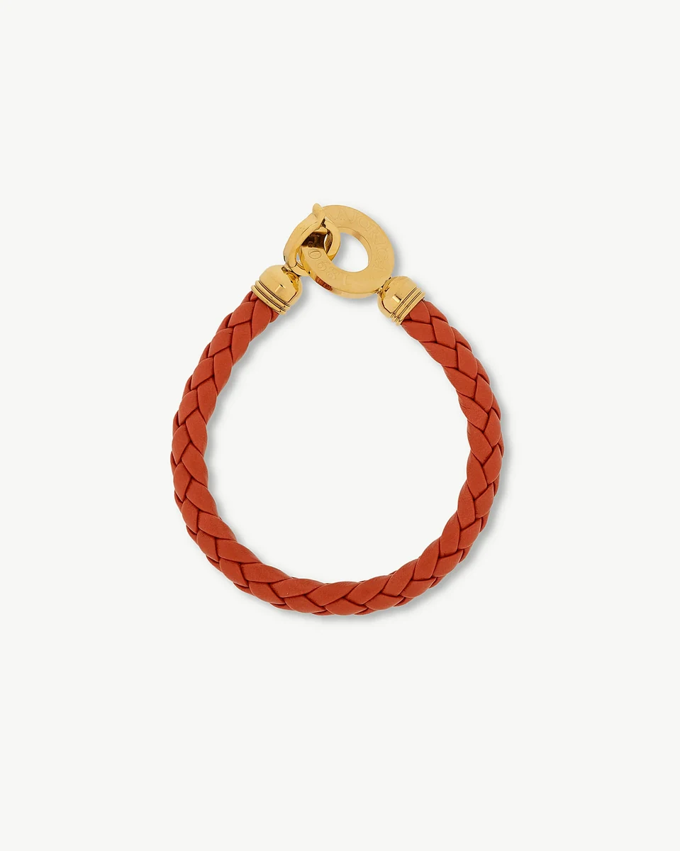 Pulsera de cuero trenzado naranja con cierre circular de acero dorado, vista frontal sobre fondo blanco.