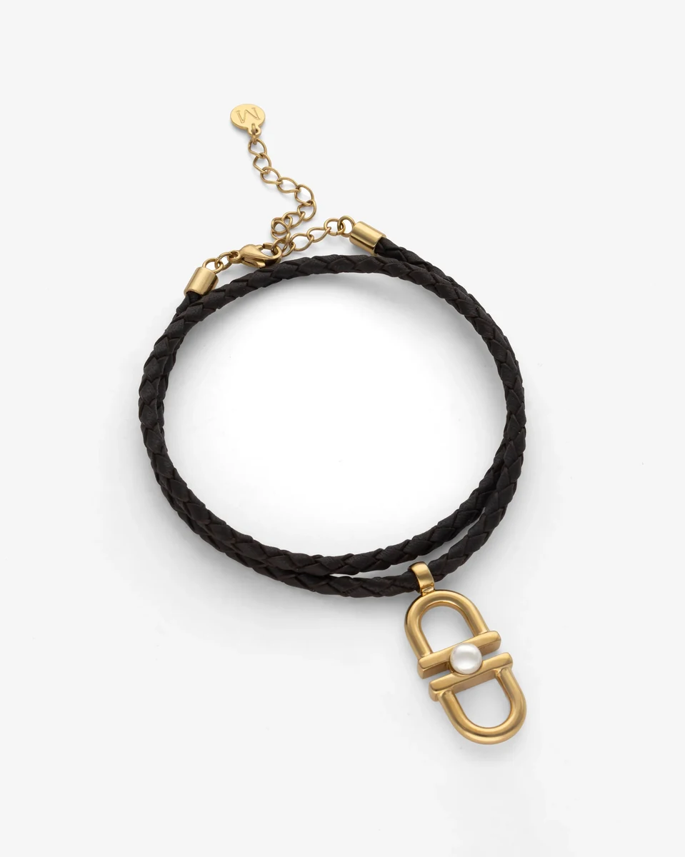 Schwarze geflochtene Lederhalskette mit goldenem Gliedanhänger und Perle, Draufsicht auf weißem Hintergrund.
