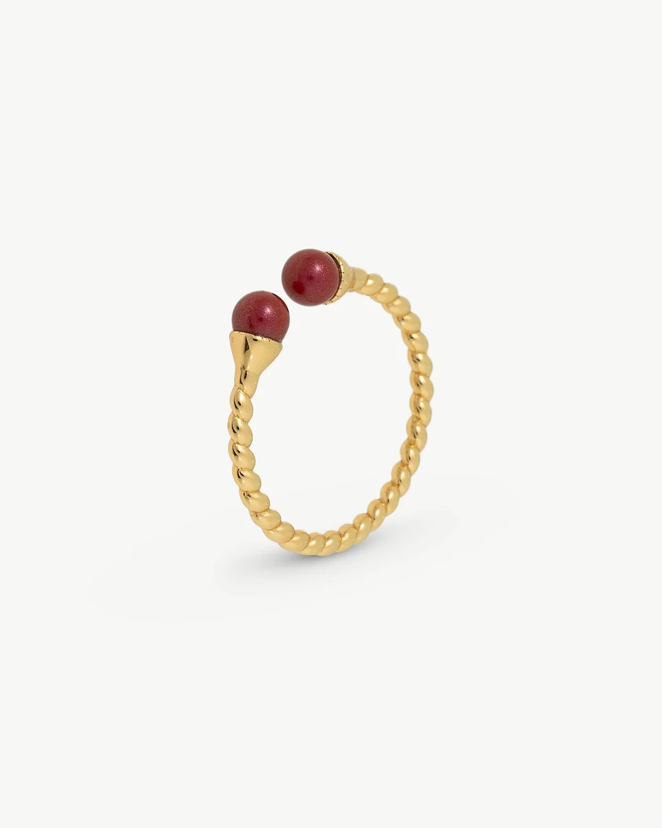 Anillo dorado con diseño trenzado y dos perlas rojas, vista lateral en ángulo sobre fondo blanco.