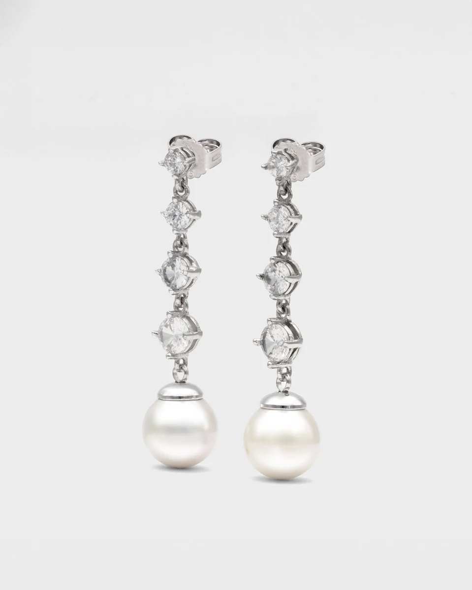Vue de face de boucles d'oreilles pendantes avec zircons et perle blanche, fond blanc neutre.