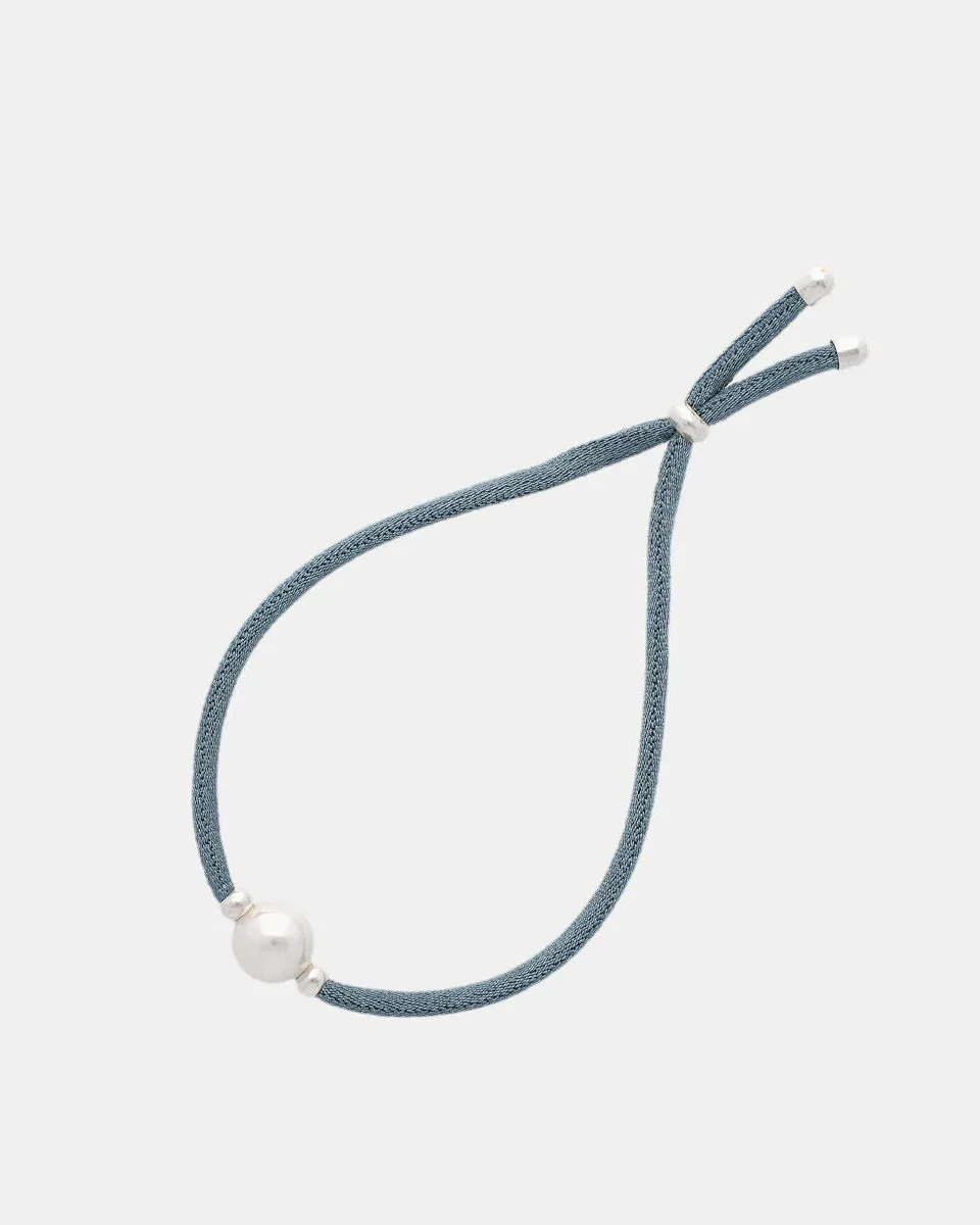 Pulsera de cordón azul con una perla blanca central, vista cenital sobre fondo gris claro.