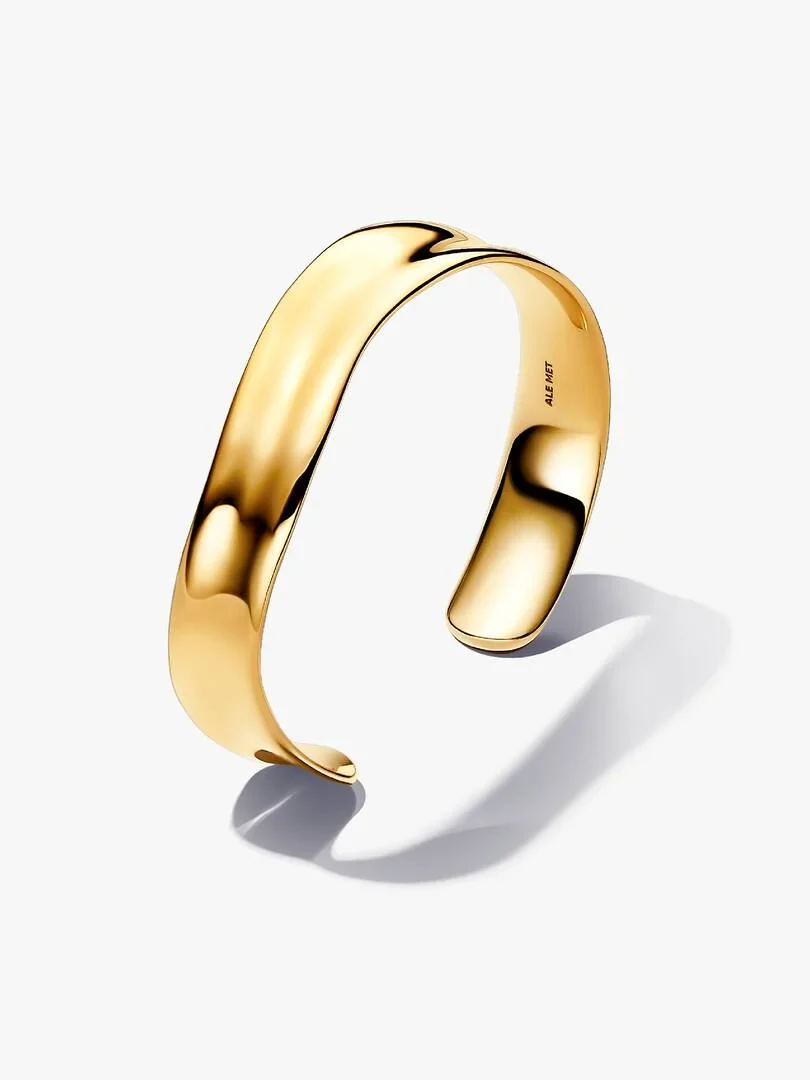 Pulsera rígida dorada con perfil ondulado, vista lateral en ángulo sobre fondo blanco con sombra suave.