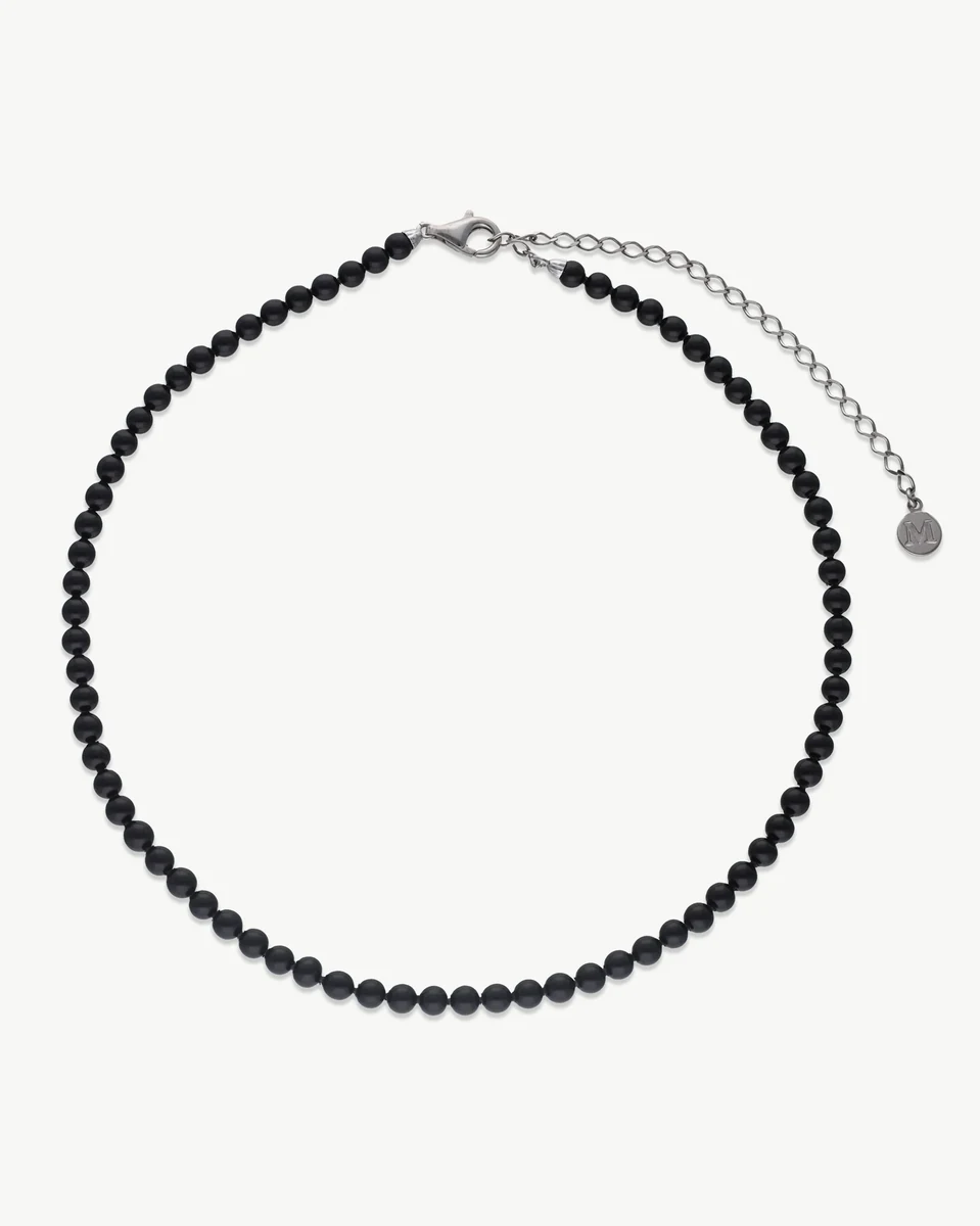 Collier de perles noires disposé en cercle sur fond blanc, vue de face avec fermoir et chaîne en argent.